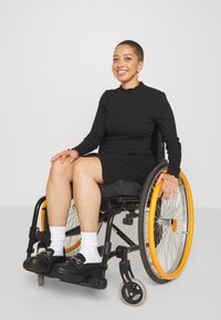 Modell i en svart klänning med långa ärmar, sittande i en rullstol med orange hjul. Hon har på sig svarta skor och vita strumpor, ler och poserar lugnt.