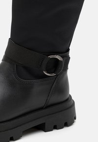 Schwarzer Knöchelstiefel mit einem glatten Lederoberteil, gummierter Sohle mit Profil und einem Textilriemen mit einem runden Metallverschlussakzent.