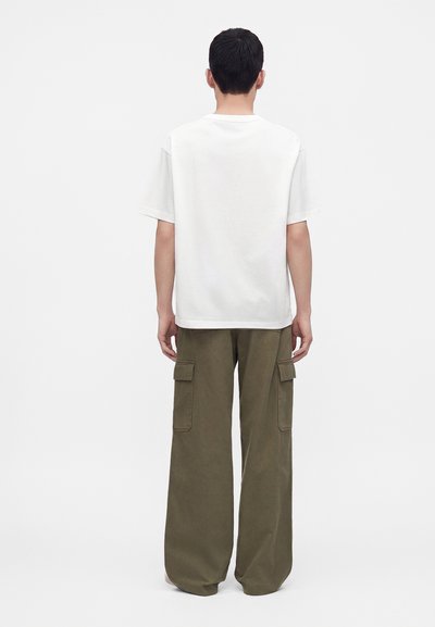 Homme debout, de dos, portant un t-shirt blanc uni à manches courtes et un pantalon cargo ample vert olive avec des poches.