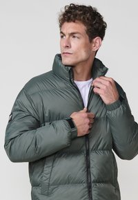 Chaqueta acolchada en verde oscuro, con un alto cuello, cierre de cremallera y tejido texturizado. Incluye puños de velcro y parche con el logo de la marca.