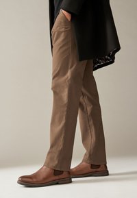 Pantalon en coton marron à coupe droite, avec poches latérales. Porté avec des bottines Chelsea en cuir marron, sur un fond neutre.