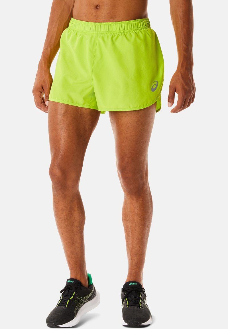 Hombre vistiendo pantalones cortos de correr de neón verde y zapatos para correr negros con detalles verdes y blancos, de pie contra un fondo liso.