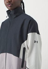 Zip-Jacke mit hohem Kragen, ausgestattet mit einem Farbkombinationsdesign in Schwarz, Weiß und Hellgrau. Enthält eine seitliche Tasche und ein Logodetail.