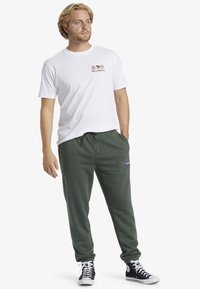 Witte crewneck t-shirt met logo op de borst, gecombineerd met donkergroene trainingsbroek met geborduurd logo, aangevuld met zwarte hoogtop sneakers.