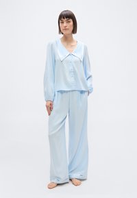 Γυναίκα όρθια με γυμνά πόδια, φορώντας ανοιχτό μπλε, φαρδιά πιτζάμα με πλατύ γιακά και ασημένιες λεπτομέρειες στον γιακά και στα τελειώματα του παντελονιού, με τα χέρια στις τσέπες.