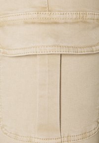 Beige kangas na zenye texture laini, ikionesha stitching inayonekana. Muundo una jaketi na mfuko wa flap na seam wima.