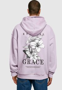 Lila Kapuzenpullover mit einem großen schwarz-weißen Blumenmuster, das Lilien und einen Schmetterling auf dem Rücken zeigt. Der Text lautet "EWIGE GNADE."