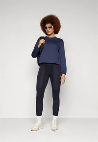 Μπλε ναυτικό αθλητικό τοπ με υφασμάτινη υφή, συνδυασμένο με μαύρα κολάν που διαθέτουν τσέπες με πατς και λείο φινίρισμα. Λευκά αθλητικά παπούτσια ολοκληρώνουν την εμφάνιση.