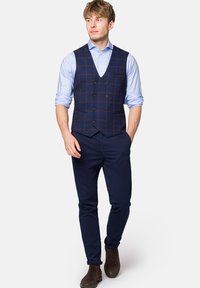 Marynarski kamizelka w kratę noszona na jasnoniebieskiej koszuli, w zestawieniu z ciemnymi spodniami i brązowymi butami. Posiada guziki i dopasowany krój.