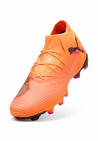 Kirkkaan oranssi jalkapallokengän teksturoitu päällinen, musta logo ja liukuvärjätty ulkopohja, jossa vaaleanpunaisia yksityiskohtia. Tyylikäs muotoilu ja tukeva istuvuus.