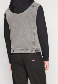 En person som bär en svart hoodie och en ljusgrå ärmlös denimjacka, med svarta byxor som har en liten logotyp på bakfickan.