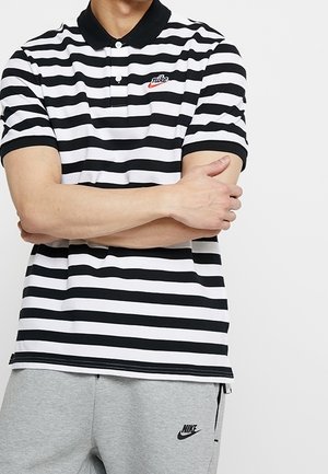 Mężczyzna noszący czarno-białą pasiastą koszulkę polo Nike z założonymi skrzyżowanymi rękami i jasnoszare spodnie dresowe Nike.