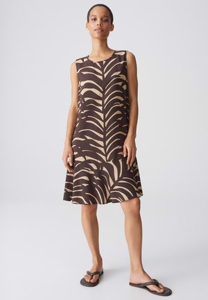 Donna in piedi con vestito senza maniche al ginocchio con motivo astratto di foglie beige e marrone scuro e infradito marrone scuro.