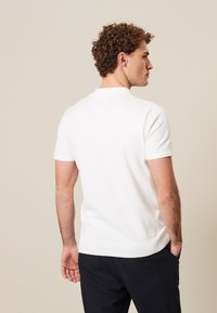 Camisa polo blanca de manga corta con cuello redondo. Presenta una textura suave y un dobladillo acanalado. El diseño es sencillo, sin patrones visibles.