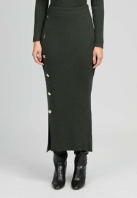 Jupe tricotée côtelée vert foncé avec des accents de boutons sur le côté, dotée d'une fente et d'une coupe ajustée, associée à des bottines noires.