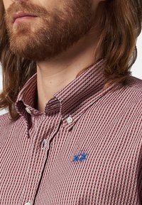 Camisa de cuadros rojos y blancos de botones con una textura suave, que cuenta con un logo bordado en azul en el pecho y un diseño con cuello.