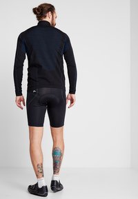 T-shirt de sport noir à manches longues avec des panneaux texturés, associé à des shorts de compression noirs. Présente des accents bleus et des tatouages visibles sur les jambes.