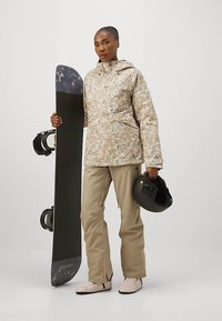 Beige kamuflaažjaketiga ja -pükstega naine hoiab ühes käes lumelauda ja teises musta kiivrit.