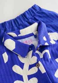 Camicia blu con bottoni, decorata con motivi astratti bianchi, texture a coste e colletto. Presenta un’etichetta con la scritta "NEXT" stampata in rosa.
