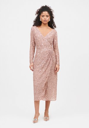 Robe mi-longue rose framboise à manches longues, design enveloppant, entièrement ornée de sequins, avec une fente sur le côté, portée avec des sandales à talons transparents.