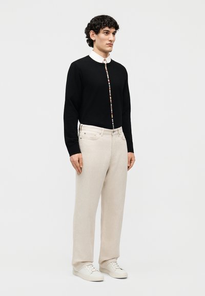 Jeune homme debout portant un pull noir avec une rayure verticale multicolore, une chemise blanche à col, un pantalon beige et des baskets blanches.