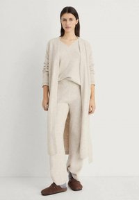 Cardigan lavorato a maglia beige e abbinato a un maglione e pantaloni, realizzati in materiale morbido. Il cardigan presenta una scollatura aperta, maniche a costine e una vestibilità comoda.