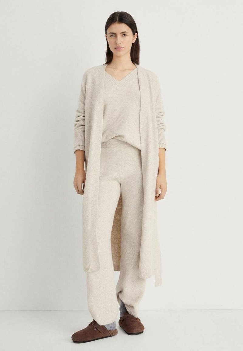 Cardigan lavorato a maglia beige e abbinato a un maglione e pantaloni, realizzati in materiale morbido. Il cardigan presenta una scollatura aperta, maniche a costine e una vestibilità comoda.