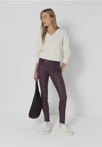 Jersey de crema con escote en V combinado con pantalones de cuero ajustados en color ciruela. La modelo sostiene un bolso de hombro marrón y lleva zapatillas blancas. Fondo sencillo.