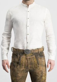 Weißes Leinenhemd mit Stehkragen, knöpfbarer Front, kombiniert mit braunen Leder-Shorts mit gelber Stickerei und einer silbernen Gürtelschnalle.