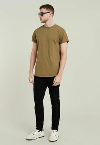 T-shirt verde oliva a maniche corte, con colletto a girocollo; abbinato a jeans neri aderenti e sneakers bianche. Modello che indossa occhiali da sole scuri.