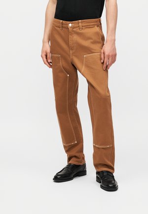 Barna munkaruhastílusú nadrág megerősített térdrésszel és fehér varrással, fekete cipővel és fekete inggel viselve.