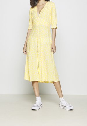 Kvinde iført en gul midi-kjole med hvidt blomsterprint, puffede ærmer, knapper foran og hvide høje sneakers.