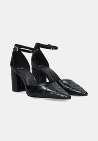 Tacones de charol negro con punta afilada, patrón texturizado de cocodrilo y tacones de bloque robustos. Correas ajustables en el tobillo con herrajes dorados.