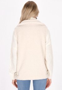 Cremfarbener Fleece-Jacke mit Zopfmusterärmeln, hohem Kragen und seitlichen Verschlüssen. Weiche Textur, lässige Passform und geschichtetes Design.