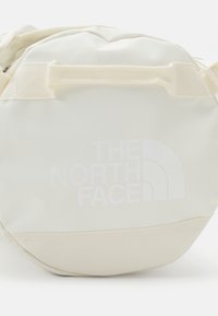 Sac bandoulière rond compact de couleur crème avec une poignée supérieure et le logo blanc "THE NORTH FACE" à l'avant.