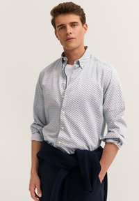 Mann trägt ein weißes gemustertes Hemd über einem weißen T-Shirt, mit einem marineblauen Pullover um die Taille gebunden, vor einem schlichten Hintergrund.