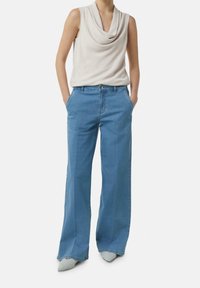 Femme portant un haut crème sans manches drapé, un jean large bleu clair et des chaussures pointues de couleur claire, mains dans les poches, debout sur fond blanc.