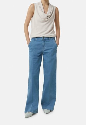 Žena v bezrukávovém krémovém topu s volnými prvky, světle modrých širokých džínách a světlých špičatých botách, s rukama v kapsách, stojící na bílém pozadí.