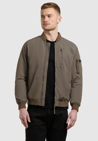 Homme portant une veste aviateur marron avec des poches zippées sur la poitrine et les manches, debout devant un fond gris clair uni.