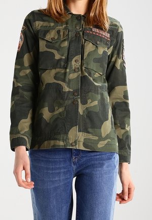 Vrouw draagt een groen-bruine camouflagejas met meerdere voorzakken en emblemen, gecombineerd met een blauwe spijkerbroek.