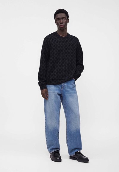 Jeune homme debout portant une chemise noire à motifs à manches longues, un jean large bleu clair et des chaussures noires sur fond blanc.