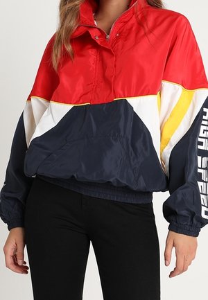 Vrouw die een colorblock windjack draagt met rode, marineblauwe, witte en gele delen en tekst op de linker mouw, gecombineerd met een zwarte broek.