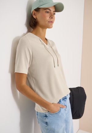 Jonge vrouw met een beige korte mouw hoodie, lichtblauwe spijkerbroek en een lichtgroene pet, leunend tegen een witte muur.