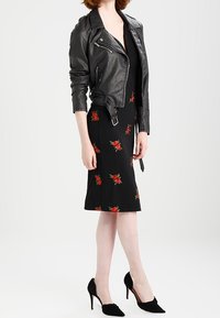 Kobieta w czarnej skórzanej kurtce nosząca czarną sukienkę do kolan w czerwone róże oraz czarne szpilki, stojąca na jednolitym tle.