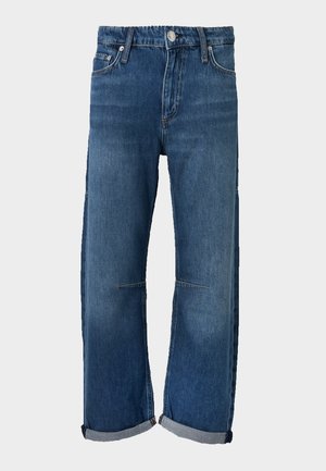 Femme portant une chemise blanche à manches longues avec col, un jean bleu retroussé et des chaussures à talons hauts noires à bouts ouverts, debout devant un fond uni.