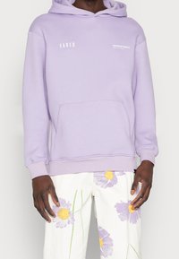 Felpa lilla chiara con tasca a marsupio, polsini a coste e un logo bianco. Abbinata a pantaloni bianchi con stampe floreali colorate.