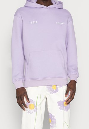 Felpa lilla chiara con tasca a marsupio, polsini a coste e un logo bianco. Abbinata a pantaloni bianchi con stampe floreali colorate.