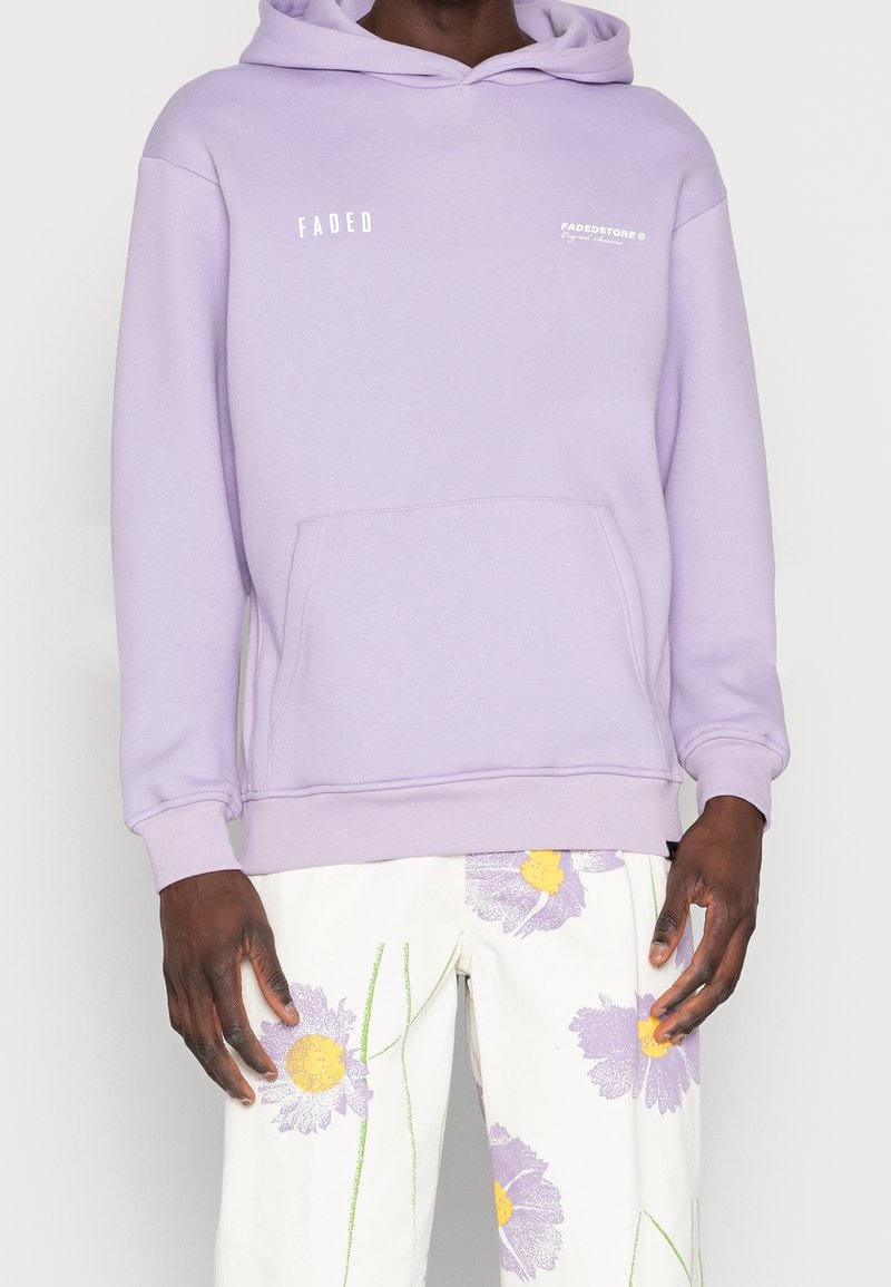 Felpa lilla chiara con tasca a marsupio, polsini a coste e un logo bianco. Abbinata a pantaloni bianchi con stampe floreali colorate.