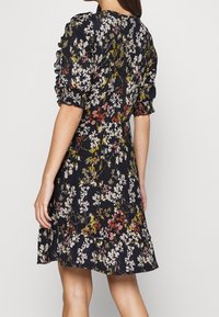 Femme portant une robe noire à mi-longueur avec un motif floral multicolore et des manches bouffantes, vue de dos.