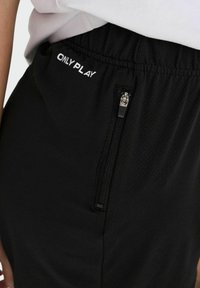 Gros plan d'un pantalon noir avec une ceinture élastique, une poche zippée verticale et le texte "ONLY PLAY" près de la taille.